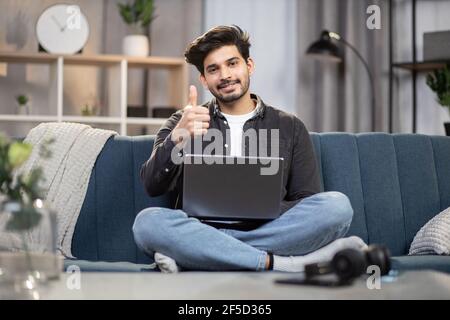 Freie Freizeit, moderner Job-Lebensstil, freiberuflich. Junge entspannte sich gut gelaunt, bärtiger Inder, saß zu Hause auf dem Sofa, arbeitete online am Laptop, benutzte das Internet, lächelte und zeigte den Daumen hoch. Stockfoto