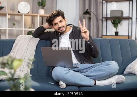 Konzept von jungen Menschen, die zu Hause mobile Geräte nutzen. Lächelnder bärtiger arabischer Inder, der zu Hause am Laptop arbeitet, während er auf dem Sofa sitzt und der Kamera ein Siegeszeichen zeigt. Stockfoto