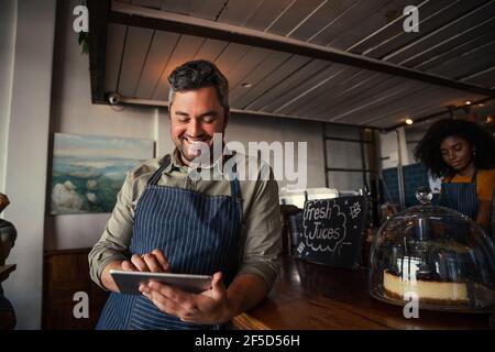 Männliche Kellner Browsen auf digitalen Tablet Scrollen durch Menü stehend Gegen die Theke im funky Cafe Stockfoto