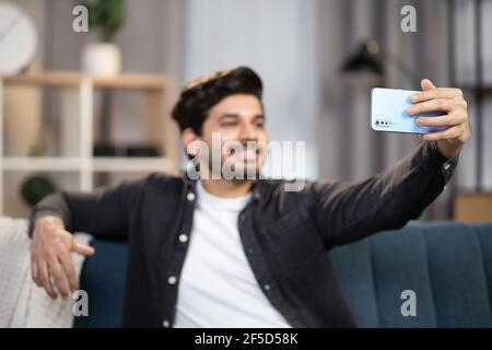 Videoanruf, Ferngespräch, Konzept sozialer Netzwerke. Verschwommenes Bild eines jungen Arabers, der zu Hause auf dem Sofa sitzt, per Smartphone Videoanruf abruft, redet und Spaß hat. Konzentrieren Sie sich auf das Telefon. Stockfoto