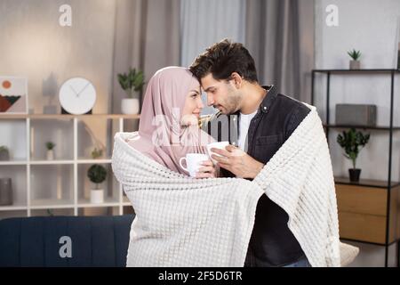 Nahaufnahme eines anhänglichen jungen arabischen Paares, das sich zu Hause unter der Decke umarmt und sich gegenseitig ansieht, die Stirn berührt, Kaffee oder Tee trinkt. Stockfoto