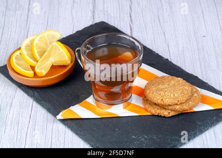 Haferflockenkekse auf Serviette und rechteckigem Steinteller, begleitet von einer Tasse Tee und einer Untertasse mit Zitronenscheiben Stockfoto