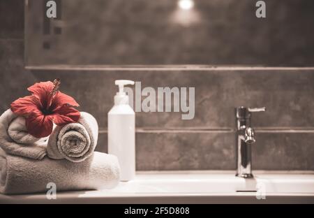Spa-Komposition mit gerollten sauberen Handtüchern, frischen Blumen und einem Pflegeprodukt in einer Flasche mit Spender am Waschbecken im Badezimmer. Stockfoto