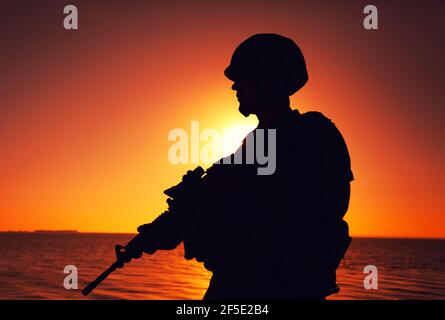 Silhouette der Armee Infanterie Soldat, Spezialeinheiten Rifleman bewaffnet mit Service Gewehr, patrouillierende Küste bei Sonnenuntergang. Küstenwache Kämpfer in Kampfhelm und taktische Munition stehen am Meer Stockfoto