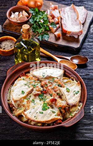Tartiflette, French Potato, Bacon und Käseauflauf in einer Tonschale auf einem dunklen Holztisch mit Löffeln, vertikaler Blick, französische Küche Stockfoto