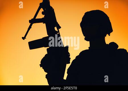 Schulter Silhouette der Armee Maschinenpistole, spezielle Operationen Kräfte Infanteristen stehend mit erhobener Waffe auf dem Hintergrund des Sonnenuntergangs Himmel. Marine Corps Shooter im Kampf Helm hält Waffe bei Sonnenaufgang Stockfoto