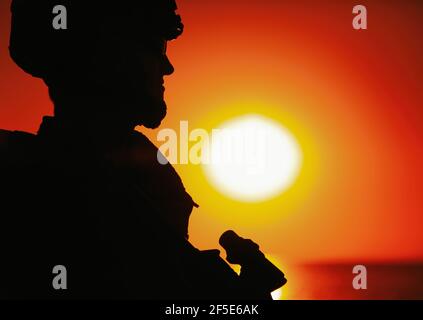 Schulter Silhouette der Armee Soldat, spezielle Operationen Kräfte Kämpfer in Kampfhelm und ballistische Brille, stehend mit Pistole auf dem Hintergrund der Untersetzung unter Ozean Horizont Sonne Stockfoto