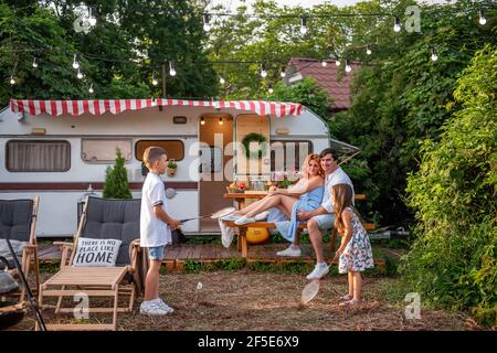 Fröhlicher Bruder und Schwester spielen Badminton durch den Anhänger LKW, glückliche Eltern sitzen auf Holzbank. Vater umarmt Mutter, Kinder haben Spaß o Stockfoto