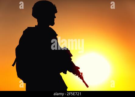 Silhouette des Armeekrieges in Helm und Munition, mit taktischem Rucksack, zu Fuß mit Service Gewehr auf dem Hintergrund des Sonnenuntergangs Himmel. Gegen terroristische Kräfte Kämpfer marschieren bei Nacht Mission Stockfoto