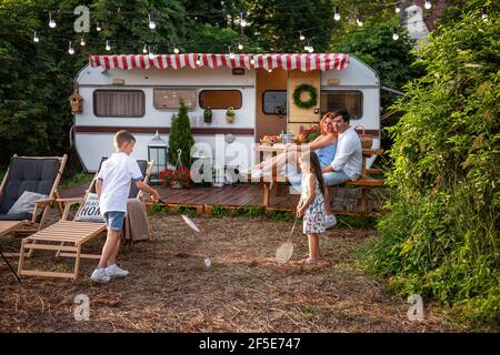 Fröhlicher Bruder und Schwester spielen Badminton durch den Anhänger LKW, glückliche Eltern sitzen auf Holzbank. Vater umarmt Mutter, Kinder haben Spaß o Stockfoto