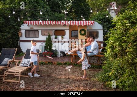 Fröhlicher Bruder und Schwester spielen Badminton durch den Anhänger LKW, glückliche Eltern sitzen auf Holzbank. Vater umarmt Mutter, Kinder haben Spaß o Stockfoto