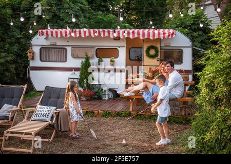 Fröhlicher Bruder und Schwester spielen Badminton durch den Anhänger LKW, glückliche Eltern sitzen auf Holzbank. Vater umarmt Mutter, Kinder haben Spaß o Stockfoto