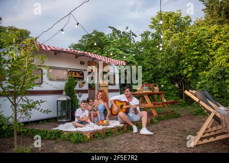 Junger Vater spielt Gitarre, rothaarige Mutter mit Kindern singt Lieder. Glückliche Familie auf Picknick, Camping mit Anhänger LKW in der Natur außerhalb der Stadt