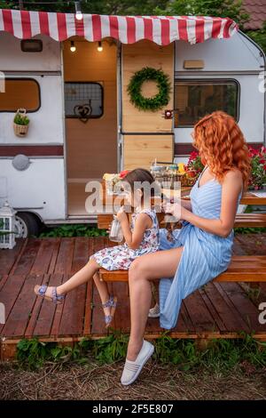 Die junge rothaarige Mutter flechtet die Zöpfe ihrer kleinen Tochter auf einer Holzbank mit einem Lastwagen. Mütterliche Versorgung. Mädchen trinkt Limonade. Picknick, Campin Stockfoto