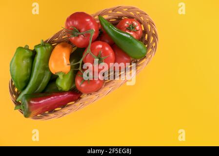 Lange Paprika und Tomaten in einem Weidenkorb. Isoliert auf gelbem Hintergrund.Kopiebereich der Draufsicht Stockfoto