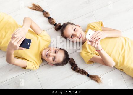 Junge Zwillingsmädchen liegen auf dem Boden, beide halten Mobiltelefone in den Händen. Stockfoto