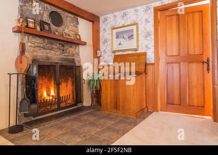 Brennter Kamin mit Feldstein Mantel-Stück und Brennholz Aufbewahrungsbox in Wohnzimmer mit beigefarbenem Teppichboden und Keramikfliesen im Inneren Ein altes Haus aus dem Jahr 1807 Stockfoto