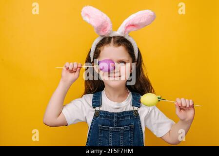Nahaufnahme Porträt von kleinen hübschen Mädchen in rosa Hase flauschige Ohren bedeckt Auge mit kleinen Ostern farbigen Eier, isoliert auf pastellgelben Studio Wand Stockfoto