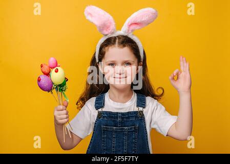 Nahaufnahme Porträt des kleinen Mädchens in Hasen flauschige Ohren halten farbige kleine Eier und zeigt okay Hand Zeichen, Modelle über gelbe Wand Stockfoto