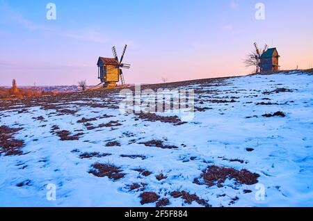 Das verschneite Feld im frühen Frühjahr mit strahlendem Sonnenuntergang Himmel darüber und die alten Holzwindmühlen am Horizont, Pyrohiv Skansen, Kiew, Ukraine Stockfoto
