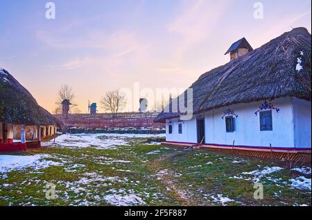 Erkunden Sie die malerischen weiß getünchten Haata-Häuser der Region Podillya mit Blick auf historische Holzwindmühlen am Sonnenuntergangshorizont, Pyrohiv Skansen, Kiew, Stockfoto