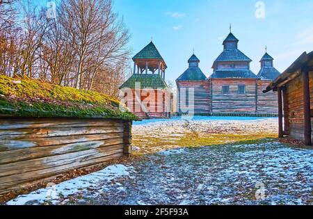 Die alte Holzkirche der Auferstehung und ihr Glockenturm mit alten Mauern der Scheunen im Vordergrund, Polissya Region Architektur, Pyrohiv Skansen, Stockfoto