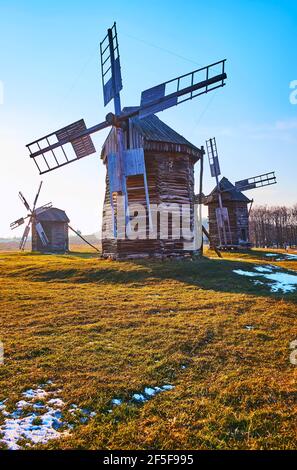 Erhaltene alte Holzwindmühlen auf der grünen Wiese mit letzten Überresten von Schnee, Pyrohiv Skansen, Kiew, Ukraine Stockfoto