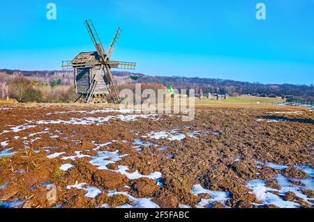 Der frühe Frühling in Pyrohiv Skansen - das gepflügte Feld ist noch mit Schnee bedeckt, und die historische Windmühle ist im Hintergrund zu sehen, Kiew, Ukraine Stockfoto
