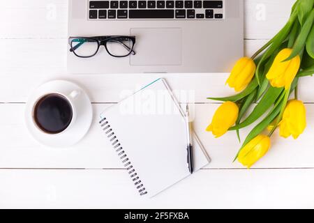 Gelbe Tulpen mit festlicher Stimmung und Kaffee auf weißen Holzbrettern. Notebook und Brille. Frauenarbeit, Gleichstellung der Geschlechter, Home Office und Karriere conce Stockfoto
