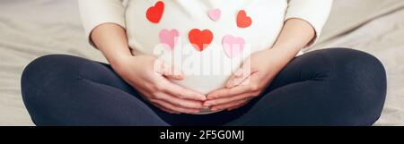 Nahaufnahme einer schwangeren Frau in weißen Kleidern, die auf dem Bett sitzen und den Bauch berühren. Schwangere Bauch mit rot rosa Papier Herzen bedeckt. Junge Dame erwartet Stockfoto
