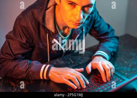 Ein männlicher Hacker tippt auf einer Laptop-Tastatur in einem dunklen Raum unter einem Neonlicht. Das Konzept von Cyberkriminalität Betrug und Identitätsdiebstahl Stockfoto