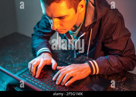Ein männlicher Hacker tippt auf einer Laptop-Tastatur in einem dunklen Raum unter einem Neonlicht. Das Konzept von Cyberkriminalität Betrug und Identitätsdiebstahl Stockfoto