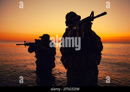 Zwei Kommandotruppen, Armee Spezialoperationsjäger zielen Sturmgewehre, während aus dem Wasser an der Küste bei Sonnenuntergang oder Morgendämmerung. Küstenwache Schützen landen an der Küste, schleichen sich im Wasser Stockfoto