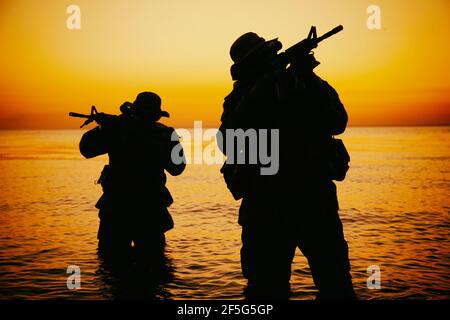 Zwei Kommandotruppen, Armee Spezialoperationsjäger zielen Sturmgewehre, während aus dem Wasser an der Küste bei Sonnenuntergang oder Morgendämmerung. Küstenwache Schützen landen an der Küste, schleichen sich im Wasser Stockfoto