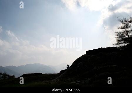 Dunkel, brüderend, dramatisch: Die Silhouette einer anonymen Figur saß auf einem Berggipfel. Scharf gegen einen hellen, wolkigen Himmel; einen Baum und weitere Berge Stockfoto