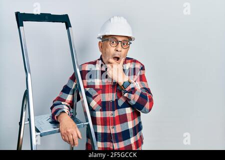 Schöner reifer Handwerker in der Nähe von Baustufen tragen Hardhat schockiert Abdeckung Mund mit Händen für Fehler. Secret Konzept. Stockfoto