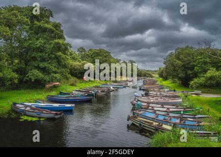 Hölzerne Paddelboote oder Motorboote liegen in Reihe auf Fluss oder Kanal umgeben von üppigen grünen Bäumen in der Nähe von Lough Leane, Ring of Kerry, Killarney, Irland Stockfoto