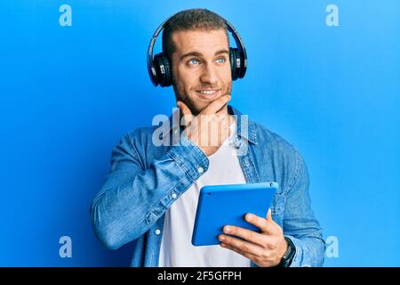 Junge kaukasische Mann mit Touchpad trägt Kopfhörer ernstes Gesicht denken über Frage mit Hand auf Kinn, nachdenklich über verwirrende Idee Stockfoto