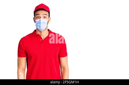 Junge gutaussehende hispanischen Mann trägt Lieferung Uniform und medizinische Maske erschrocken und erstaunt mit offenem Mund für Überraschung, Unglauben Gesicht Stockfoto
