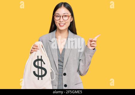 Junge chinesische Frau trägt Business-Anzug halten Dollar Tasche lächelnd Glücklich zeigen mit Hand und Finger zur Seite Stockfoto