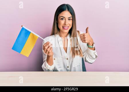 Junge hispanische Frau hält ukraine Flagge sitzen auf dem Tisch lächelnd glücklich und positiv, Daumen nach oben tun ausgezeichnet und Zustimmung Zeichen Stockfoto