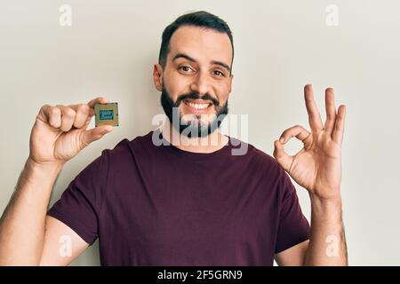 Junger Mann mit Bart hält cpu-Computer-Prozessor tun ok Zeichen mit den Fingern, lächelnd freundlich gestikulieren ausgezeichnete Symbol Stockfoto