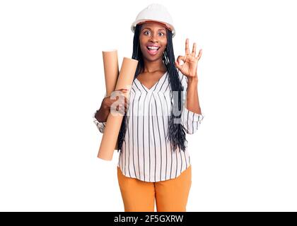Junge afroamerikanische Frau trägt Architekt Hardhat hält Blaupausen tun ok Zeichen mit Fingern, lächelnd freundlich gestikulieren ausgezeichnete Symbol Stockfoto