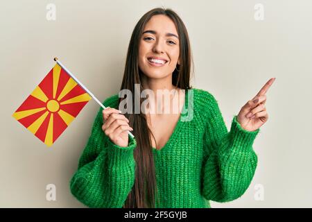 Junge hispanische Mädchen hält mazedonische Flagge lächelnd glücklich zeigt mit Hand und Finger zur Seite Stockfoto