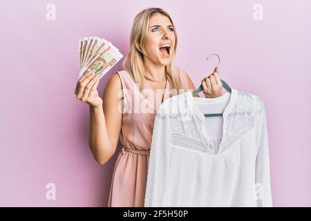 Junge kaukasische Frau hält Kleiderbügel mit T-Shirt und russischen Rubel Banknoten wütend und verrückt schreien frustriert und wütend, schreien mit Wut Blick Stockfoto