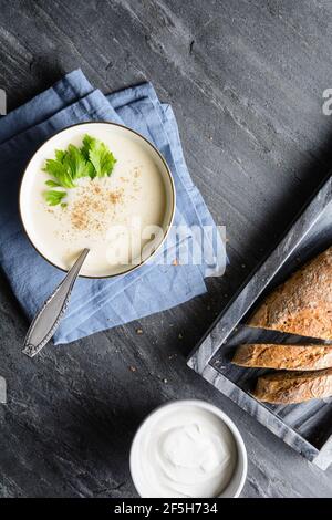 Köstliche cremige Sellerie-Suppe mit schwarzem Pfeffer und frischen Blättern, serviert mit knusprigem Brot, auf rustikalem Stein Hintergrund Stockfoto