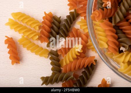 Sehen Sie durch Glasschüssel voll mit ungekochten bunten Spiralnudeln Auf Holzbrett Stockfoto