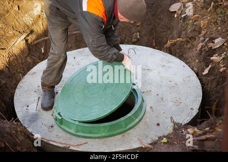 Ein Arbeiter installiert einen Kanalschacht an einem Klärbecken aus Betonringen. Bau von Kanalisationsnetzen für Landhäuser. Stockfoto