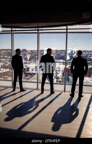 Dunkle männliche Silhouetten vor dem Hintergrund eines Panoramafensters. Männliche Geschäftsleute mit Blick auf das große Fenster eines Wolkenkratzers mit Blick auf die Metr Stockfoto