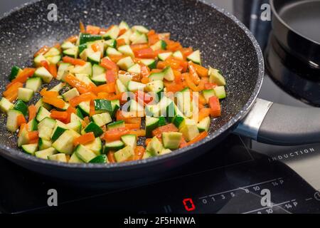 Gewürfelte Karotten, Paprika und Zucchini werden in einer schwarzen, antihaftbeschichteten Bratpfanne auf einem modernen Induktionsherd gebraten Stockfoto